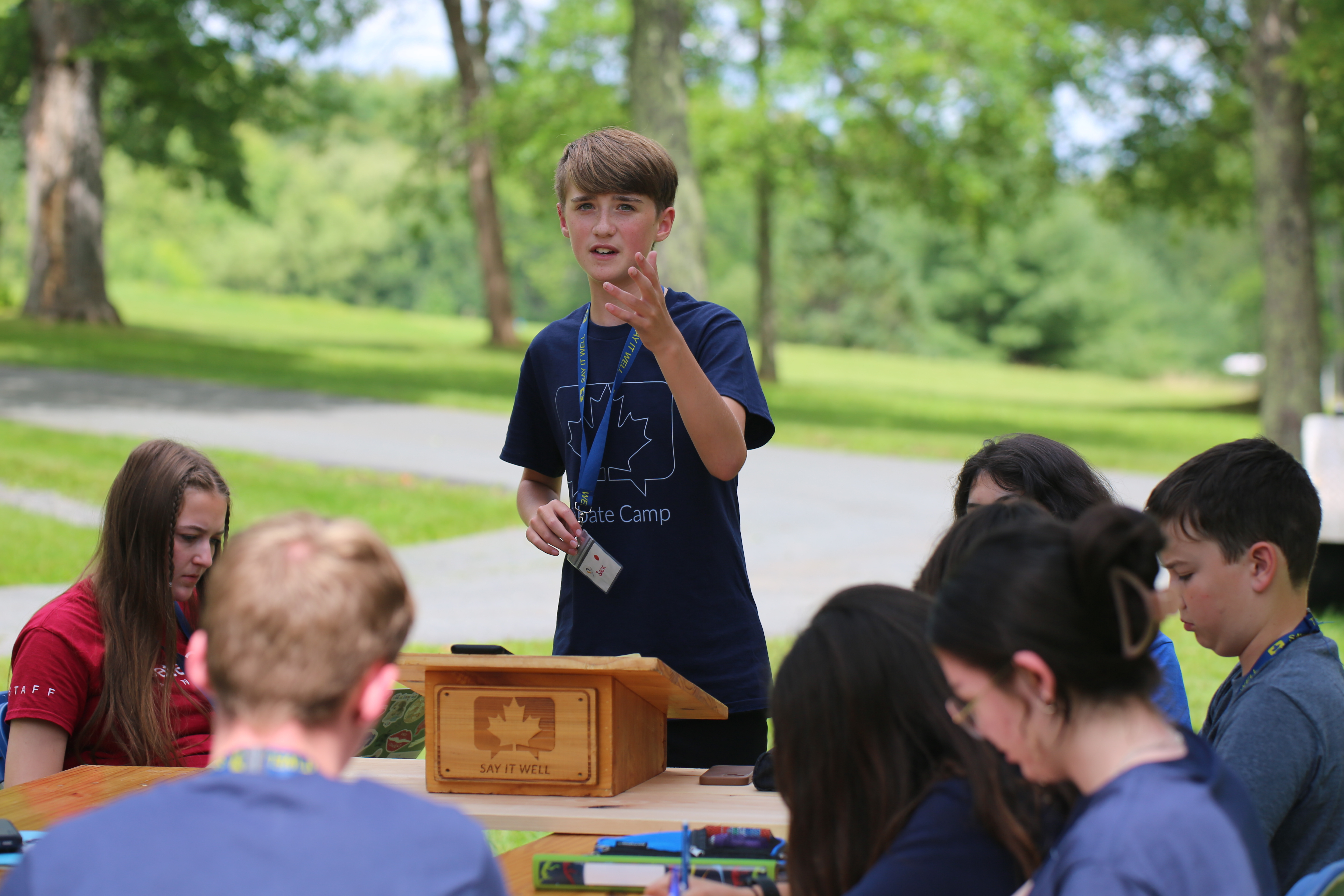 Student leading a discussion at an outdoor debate camp in Santa Barbara, speaking at a podium while other campers listen and take notes in a park-like setting